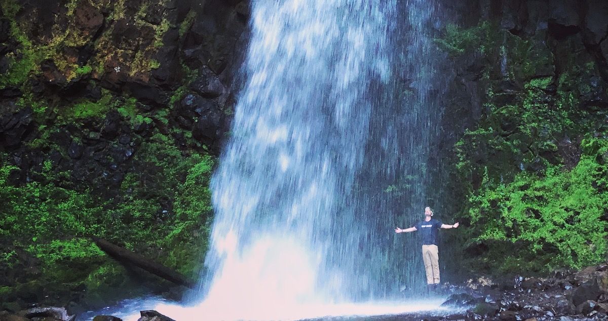 Brian Dusablon under the waterfall at Multnomah Falls in Oregon, with his arms spread wide and looking up.