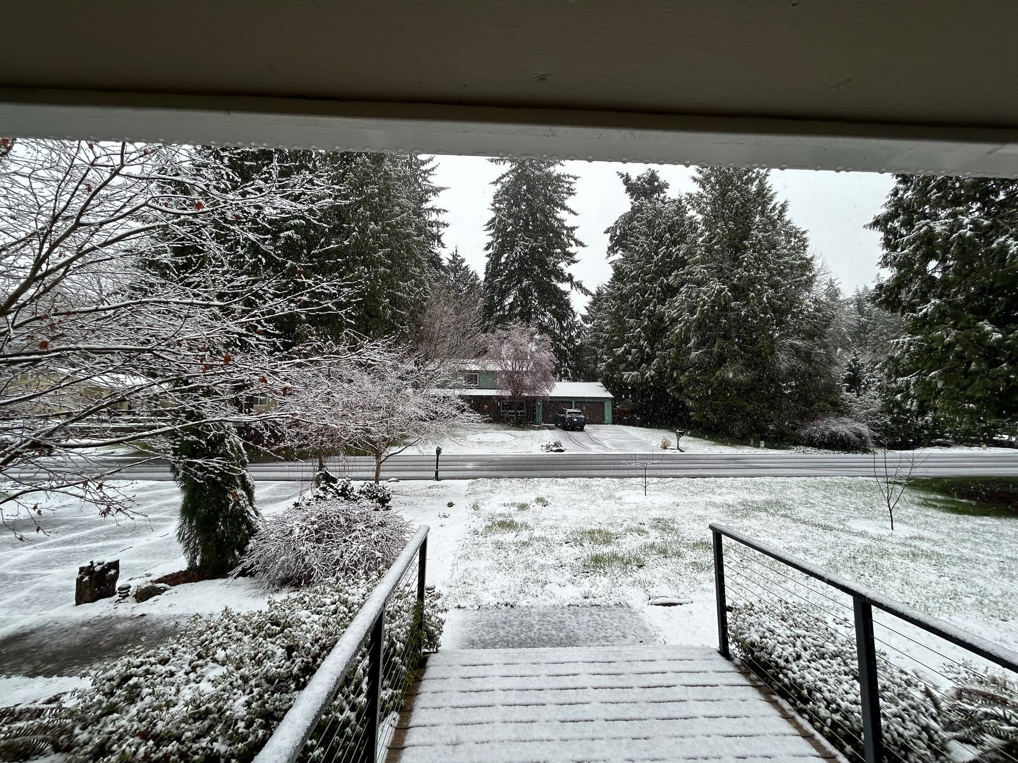 Snow covers most of the front porch of a house and the ground as you look out. They sky is gray.