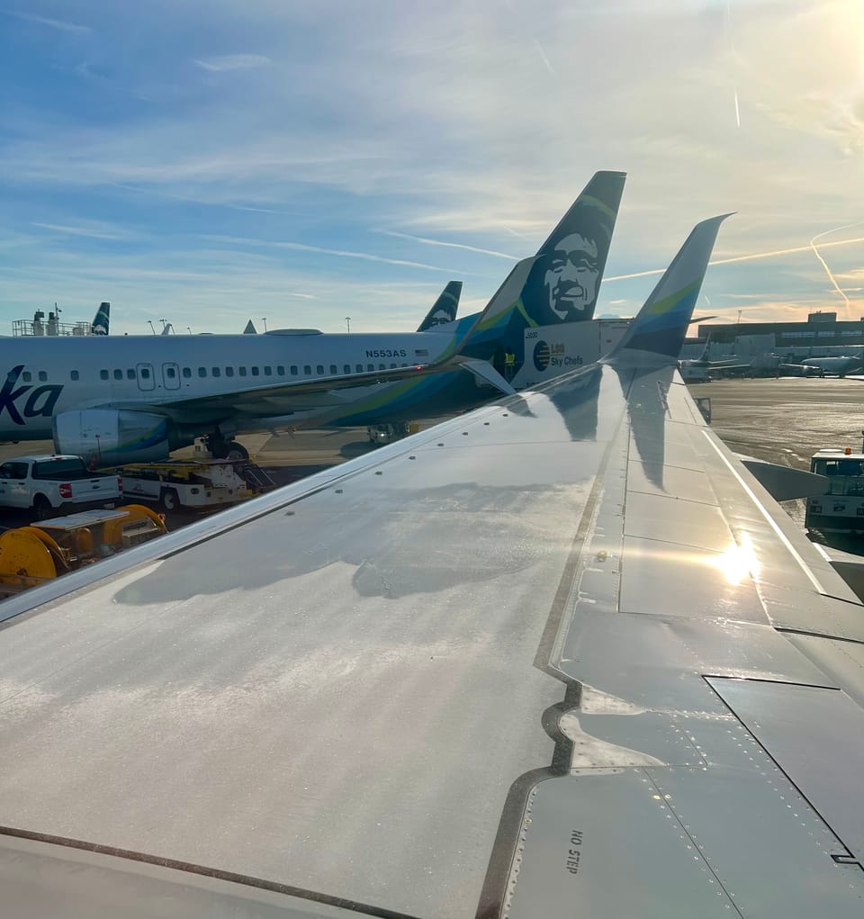 An airplane wing taken from the inside window with the sun reflecting off of it and other planes in the background.